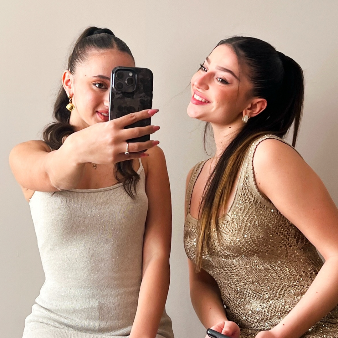 The image shows two girls in beige formal dresses taking a selfie holding a phone with designed dark floral phone case promoting the About Us page in order the people to undertstand our Shops Story.