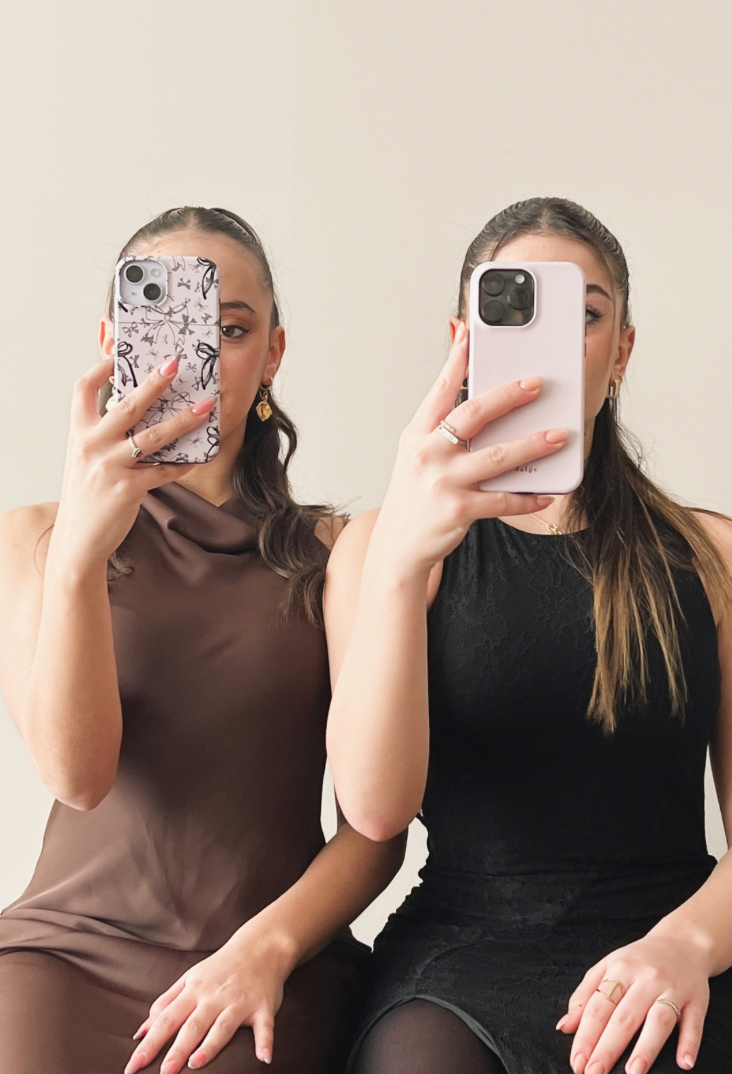 Icon featuring two girls sitting side by side, playfully holding stylish phone cases in front of their faces against a beige background, promoting the phone cases."